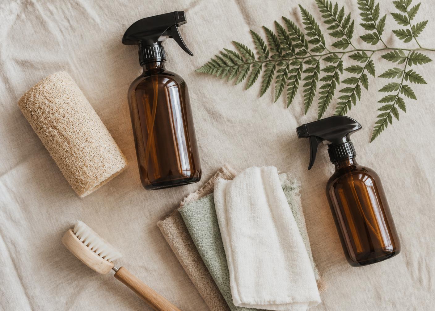 Refillable amber glass spray bottles, loofah sponge, cotton rags and a wooden brush on linen